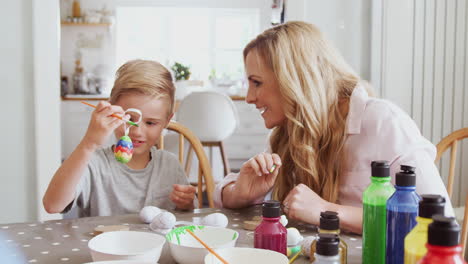mother with son sitting at table decorating eggs for easter at home
