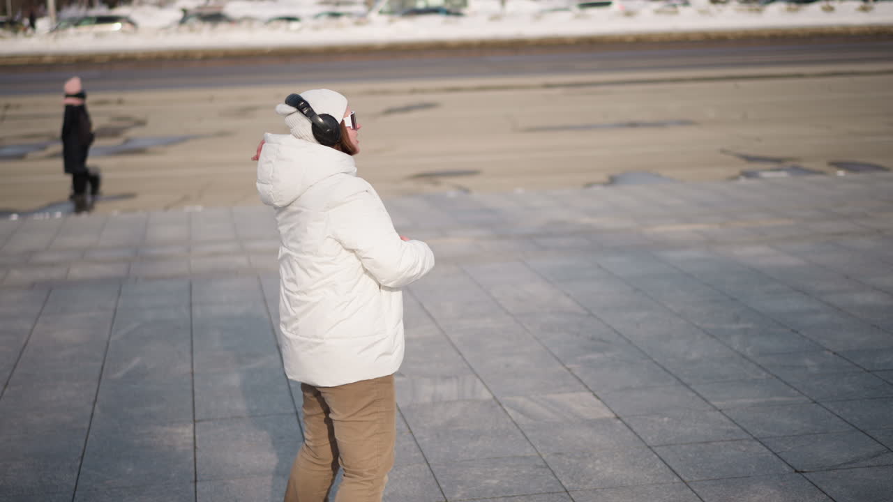 Side view of student wearing white puffer coat, beanie and headphones dancing energetically on wide tiled walkway beside busy road with passing cars and snowbanks under bright winter daylight