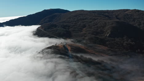 Misty-hills-covered-by-a-blanket-of-clouds-with-a-clear-blue-sky-above,-aerial-shot