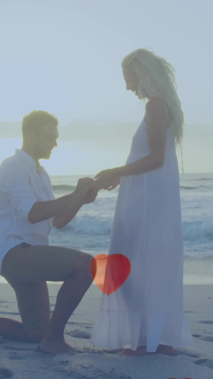 Flere hjerteballonger flytende mot en mann som frir til en kvinne med en ring på stranden.