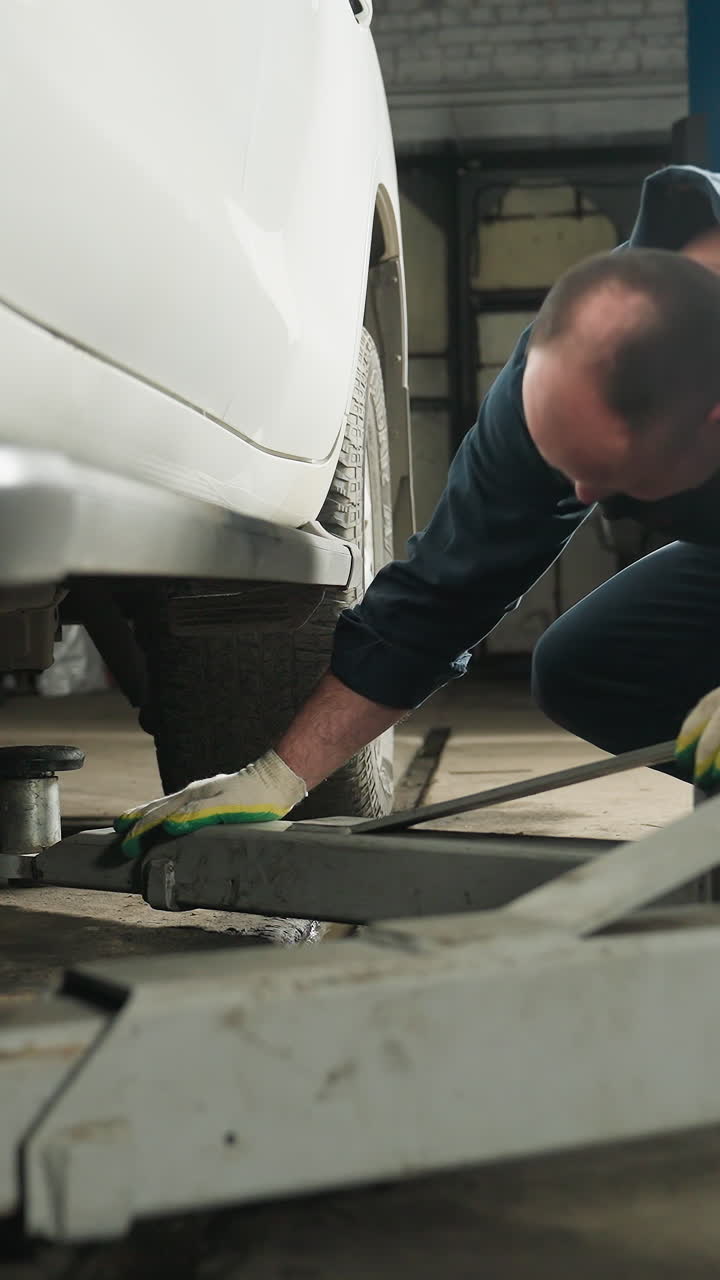 Focused view on a car jack being positioned under a vehicle in a workshop, with a mechanic's presence suggested by visible gloved hands and a blue lift pole