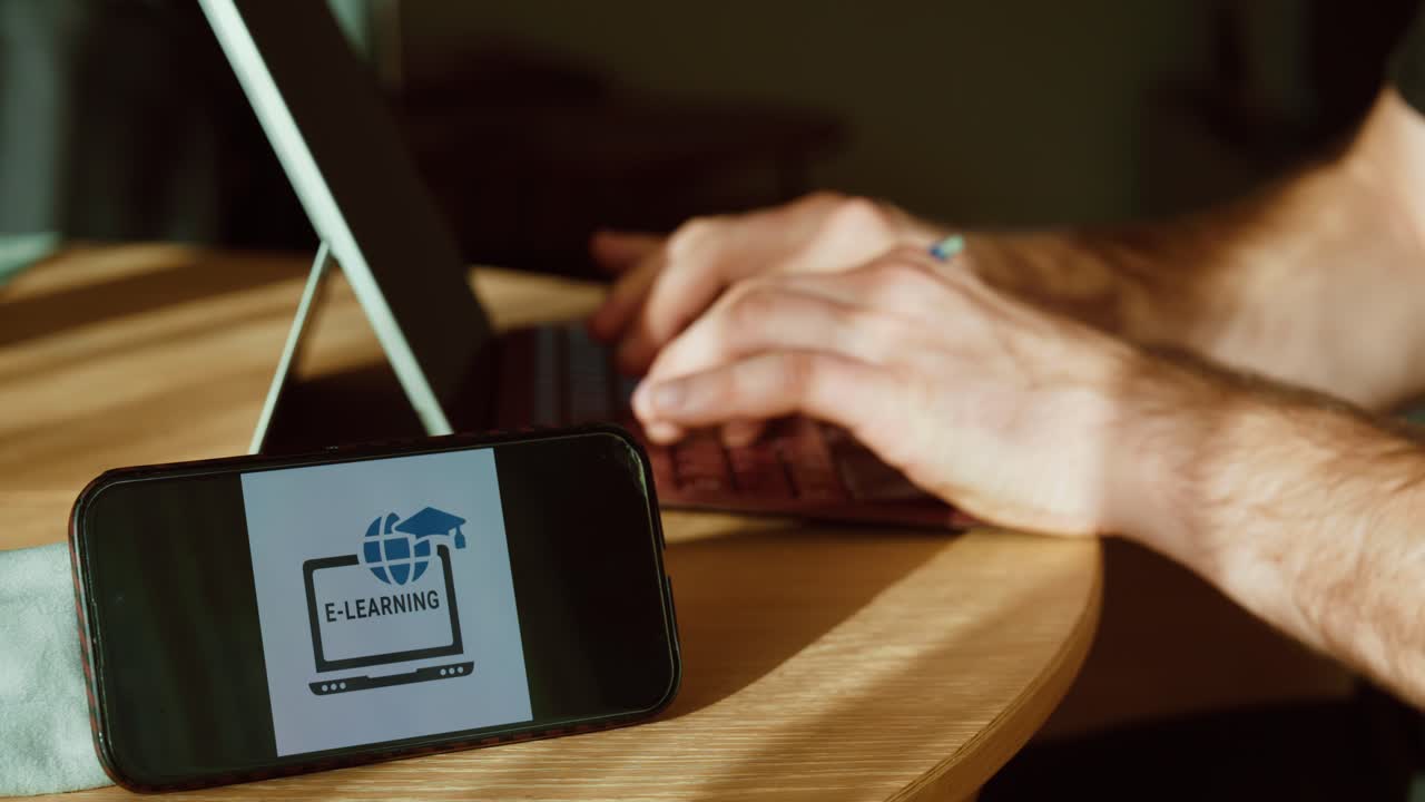 Close-up of a smartphone showing an e-learning screen next to a laptop while hands type, illustrating remote training and digital education at home or in the office. Ideal for corporate learning, comp
