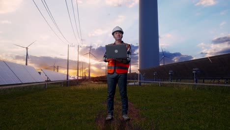 full body of asian male engineer with safety helmet working on a laptop and looking around while standing with solar panel and wind turbines