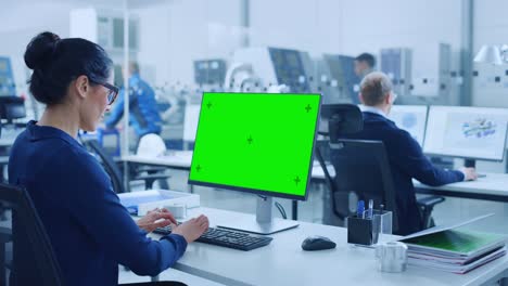 over the shoulder: female chief engineer working on personal computer, display is green mock-up screen or chroma key. industrial factory with high-tech machinery and professional workers