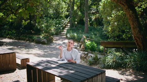 mujer sentada en la mesa del parque con un teléfono inteligente escribiendo información en un cuaderno