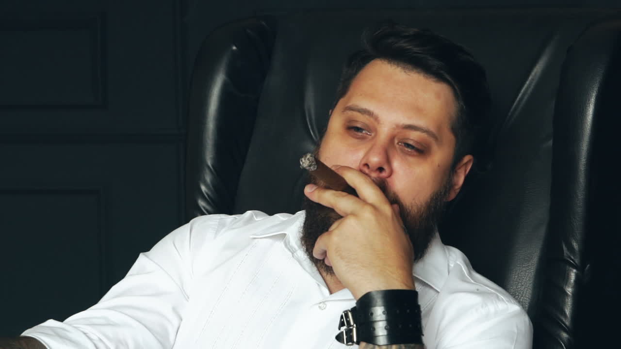 Brutal mann røyker sigar. Kjekk skjeggete mann med alvorlig ansikt holder sigar i studio