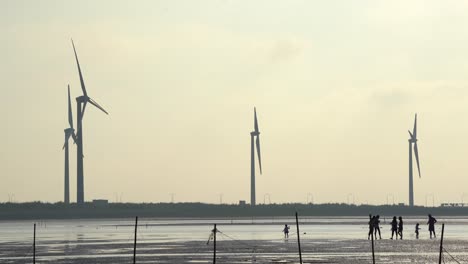 Silhouetten-Von-Touristen,-Die-Auf-Wattflächen-Spazieren-Gehen-Und-Spielen,-Mit-Sich-Drehenden-Windkraftanlagen-Im-Hintergrund,-Im-Naturschutzgebiet-Gaomei-Wetlands,-Taichung,-Taiwan