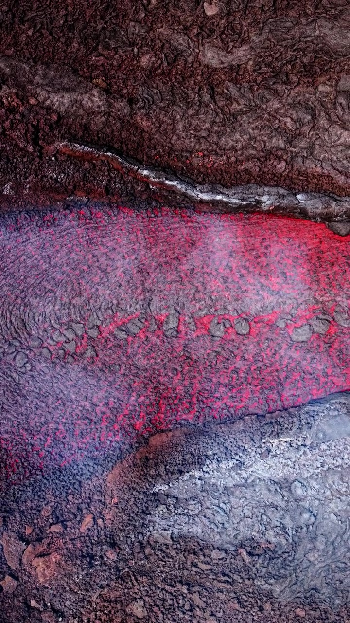 Luftfoto af glødende lava, der strømmer langs barskt vulkansk terræn med opstigende damp.
