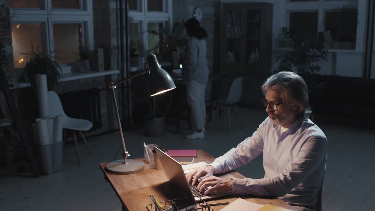 A man works late on a laptop in a modern office while a colleague moves in the background