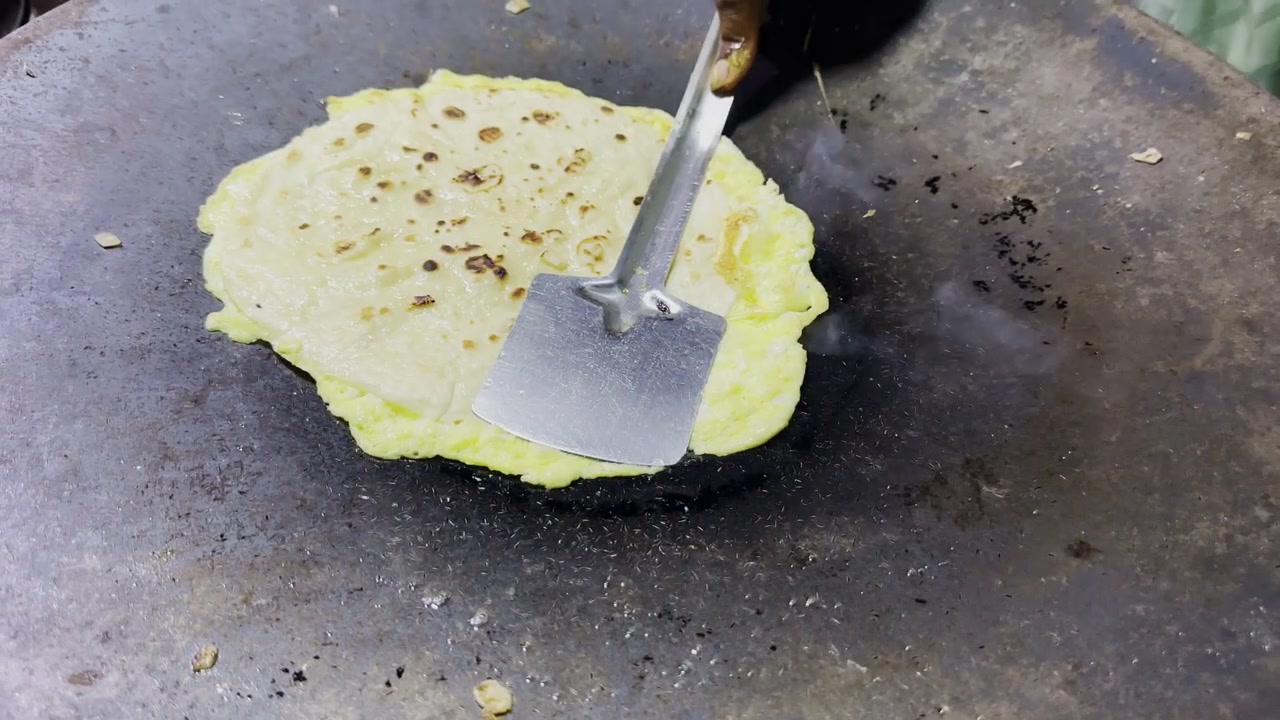 Omelette di uova in cottura su una tawa di ferro rovente, con una spatola piatta che preme la superficie, mentre si forma una crosticina dorata e si respira l'atmosfera di una tradizionale cucina di street food al chiuso!