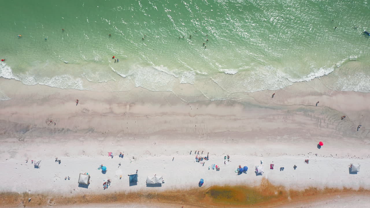 Gentle turquoise waves wash over the sandy shoreline as beachgoers relax under colorful umbrellas while swimmers play in the shallow surf from a top-down aerial perspective