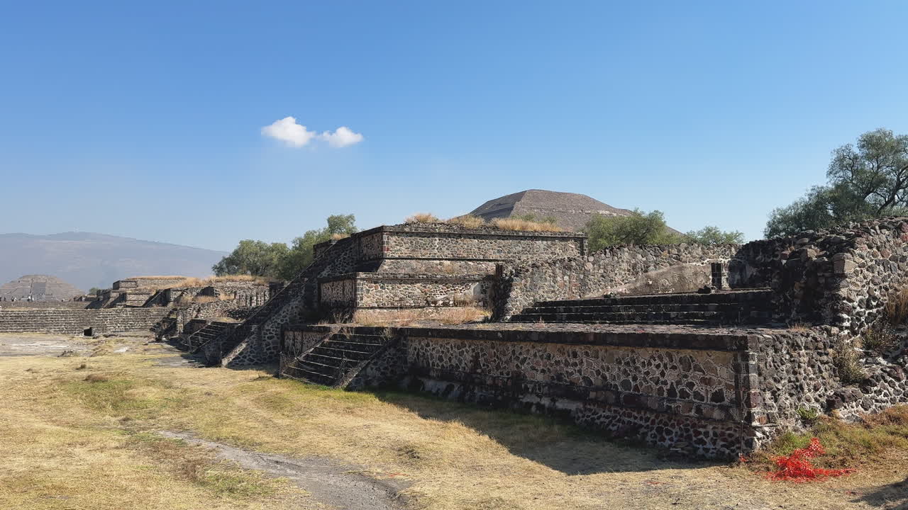 Panoramica cinematografica attraverso terrazze e scalinate in pietra di Teotihuacan con una piramide distante sotto un cielo blu, che mostra l'antica architettura messicana e filmati di repertorio di storia di viaggio.