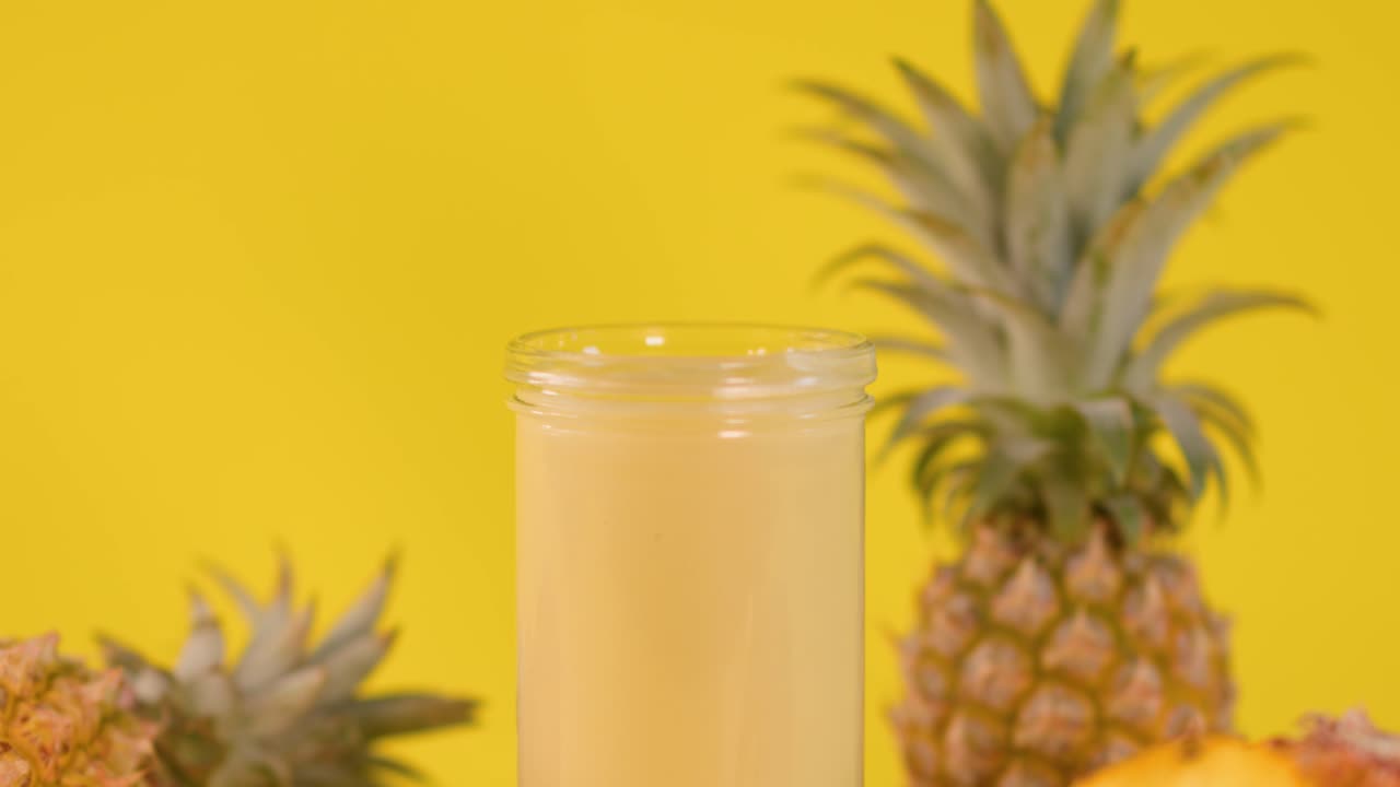 Hand places pineapple slice on creamy smoothie amid tropical fruits against a bright yellow background