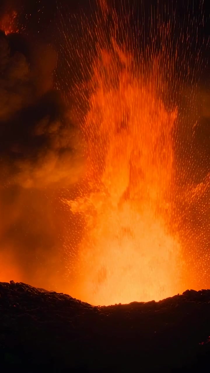 Vertikales Video: Ausbrechender Schlot stößt nachts am Kraterrand eine Lavafontäne aus, glühende Asche fällt herab.