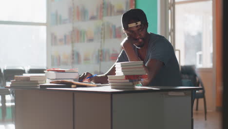 man in library sits at table with books, resting chin on hand while studying and looking thoughtful, wearing cap, teal and orange walls and shelves behind him