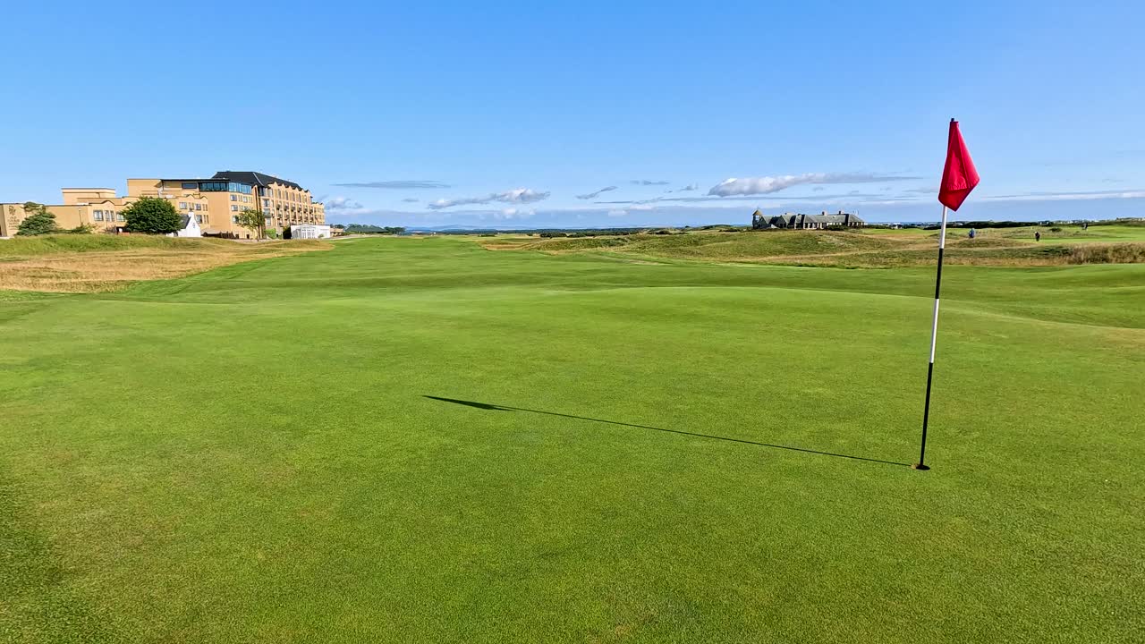 Camera slowly pans across vibrant golf green, red flag, clear sky, and distant clubhouse buildings