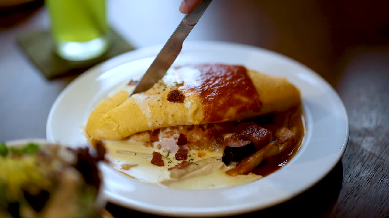 Hand slices Japanese omurice with knife, warm lighting, close-up, casual Bangkok dining setting