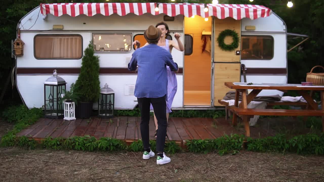 공원에서 트레일러 근처에서 즐거움을 즐기는 사랑스러운 커플, 가 ⁇ 운 소녀가 남자친구의 팔에 뛰어들어 그녀를 돌리고 있습니다. 휴가에 있는 즐거운 젊은 커플의 완벽한 사진