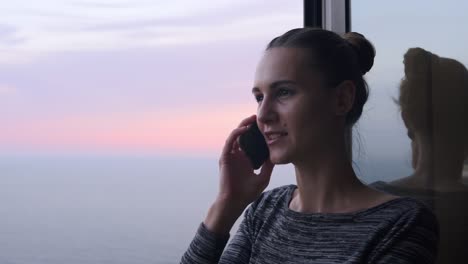 Vista-De-Cerca-De-Una-Joven-Mujer-Sonriente-Hablando-Por-Teléfono-Mientras-Está-De-Pie-Junto-A-La-Ventana-Abierta-Con-Una-Sonrisa-Durante-La-Puesta-De-Sol-Junto-Al-Mar