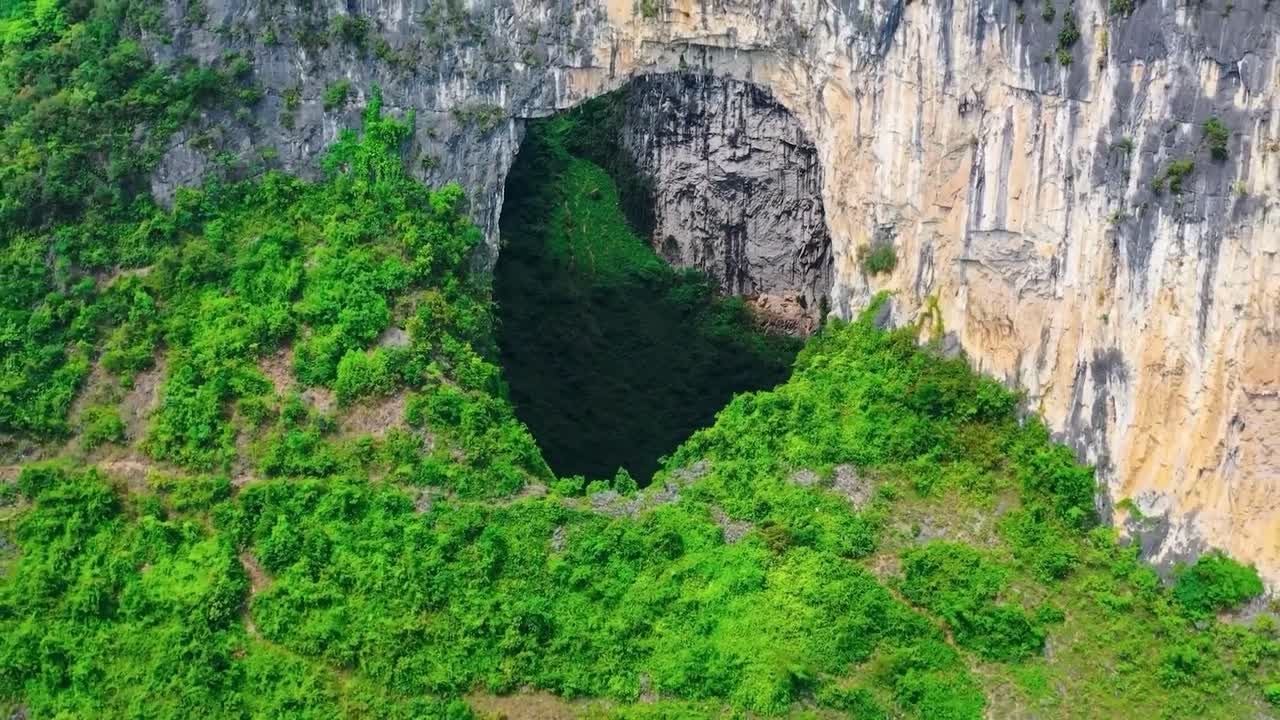 Aerial drone flying out and further into the air from Heavenly Pit of Xiaozhai Tiankeng natural sinkhole in a steep mountain cliff with green nature surrounding it in China. Sun is shining, cinematic.