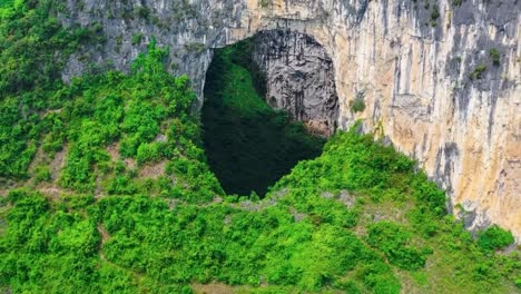 aerial drone flying out and further into the air from heavenly pit of xiaozhai tiankeng natural sinkhole in a steep mountain cliff with green nature surrounding it in china. sun is shining, cinematic.