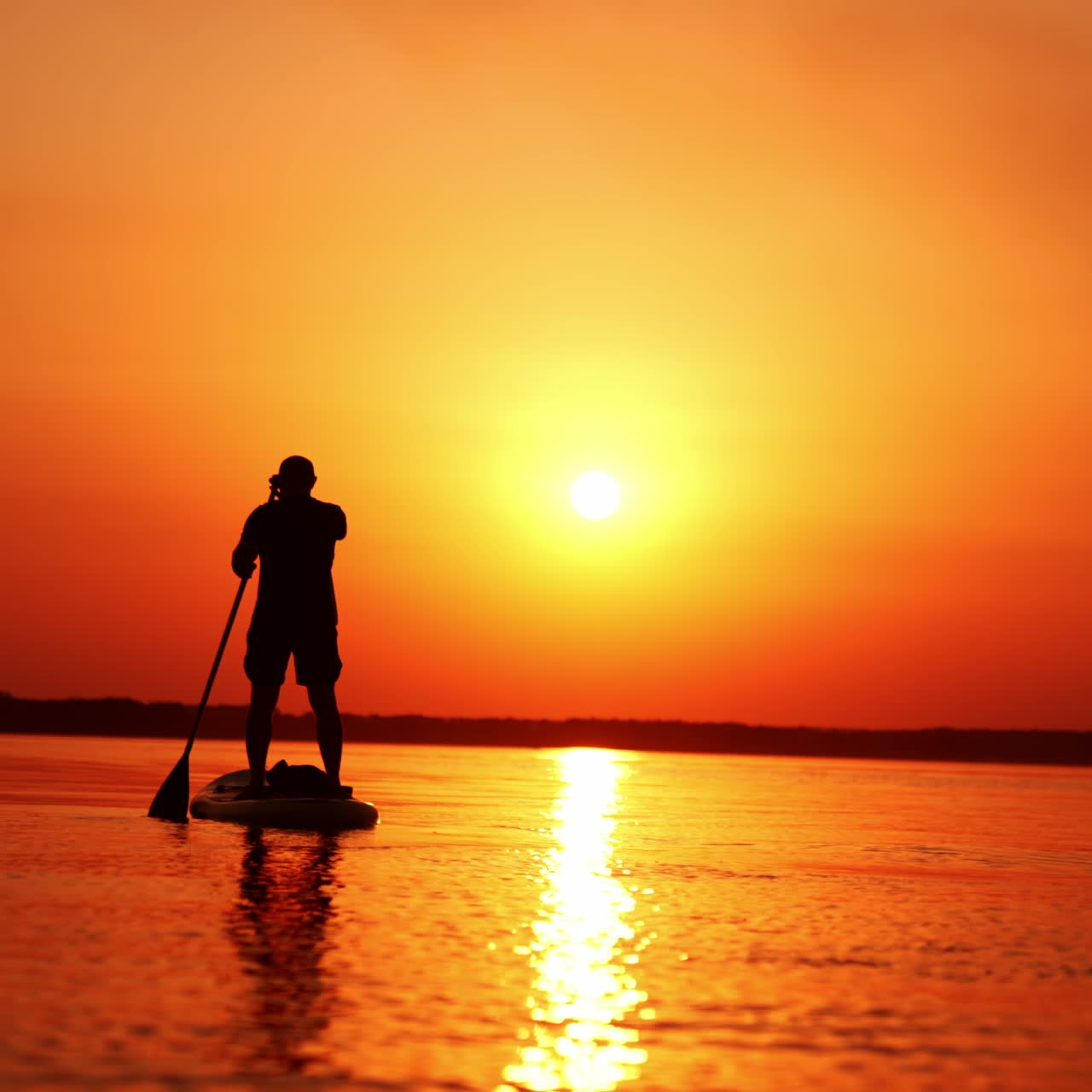 Man stands on the sup board facing the sunset. Man holds a paddle and row with it from each side