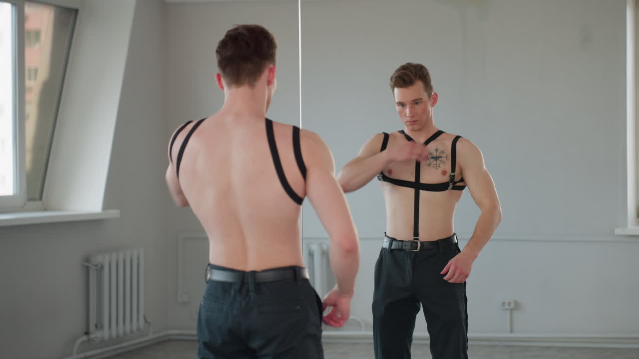 young man with defined physique adjusts harness straps around torso while standing in front of mirror in sunlit room, seen from behind with reflection showing focused expression