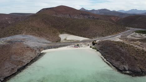 Luftschwenkaufnahme-über-Playa-El-Tecolote-In-Der-Nähe-Von-La-Paz-Baja-California-In-Mexiko-Mit-Blick-Auf-Die-Ausgetrocknete-Landschaft,-Das-Ruhige-Meer-Und-Einen-Strand-Sowie-Die-Berge-Im-Hintergrund