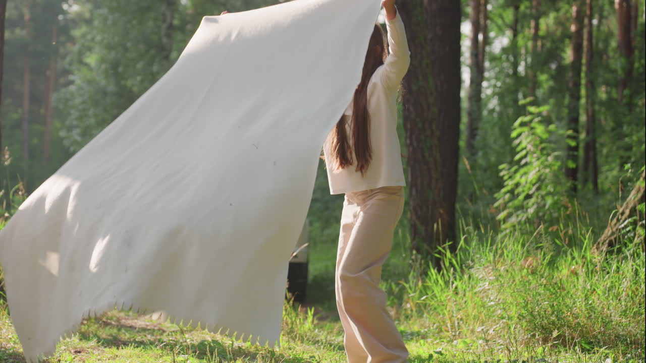 Siblings swinging white cloth playfully in forest clearing under bright sunlight, smiling and laughing together in carefree outdoor atmosphere surrounded by nature, expressing joy, and energy
