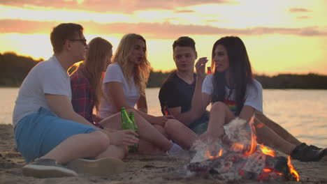 five students are sitting in shorts and t-shirts around bonfire on the sand beach. they are talking to each other and drinking beer at sunset.