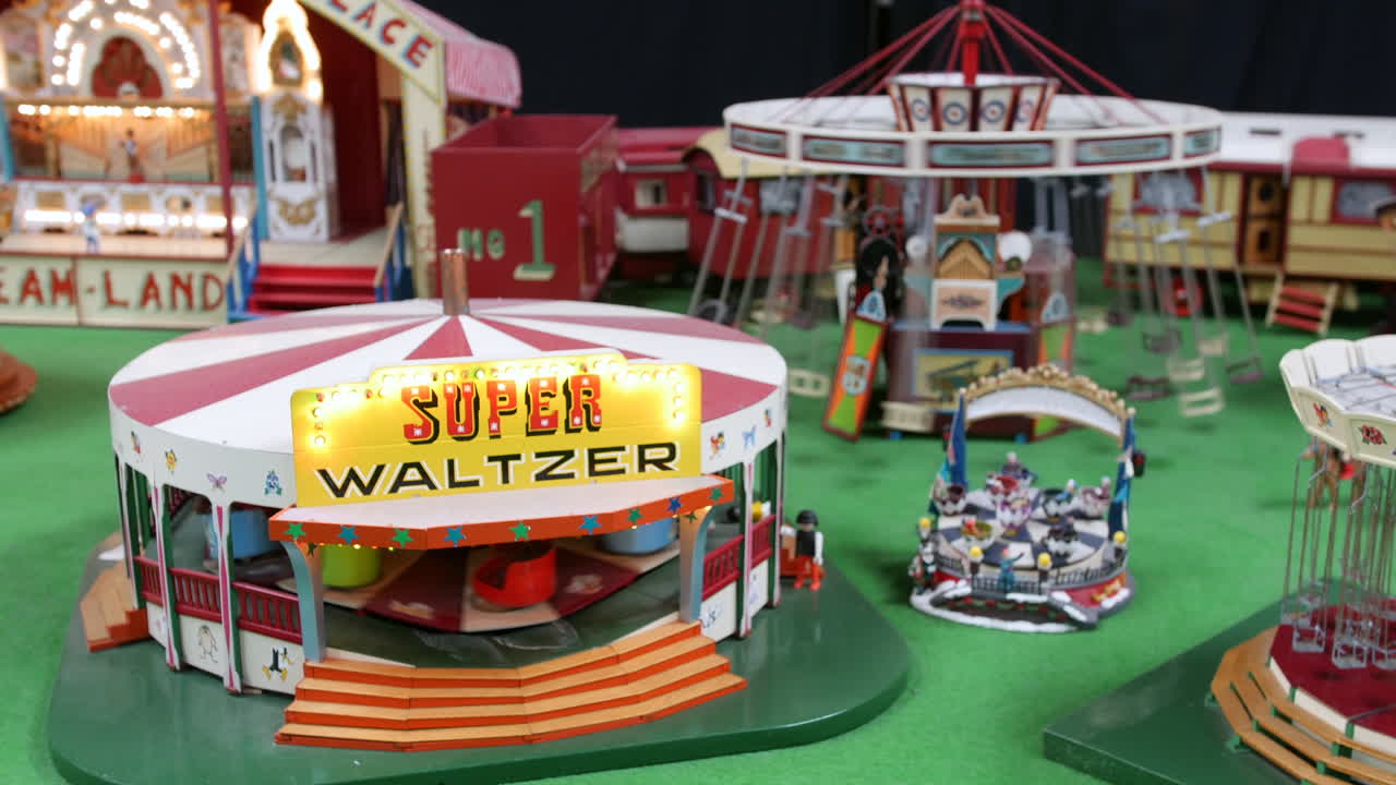 A model fairground with a Super Waltzer ride is displaying miniature amusement attractions, painted stalls, and illuminated signs on a green base