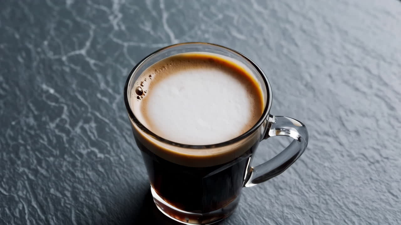 A glass mug of coffee with white foam on a dark, textured surface