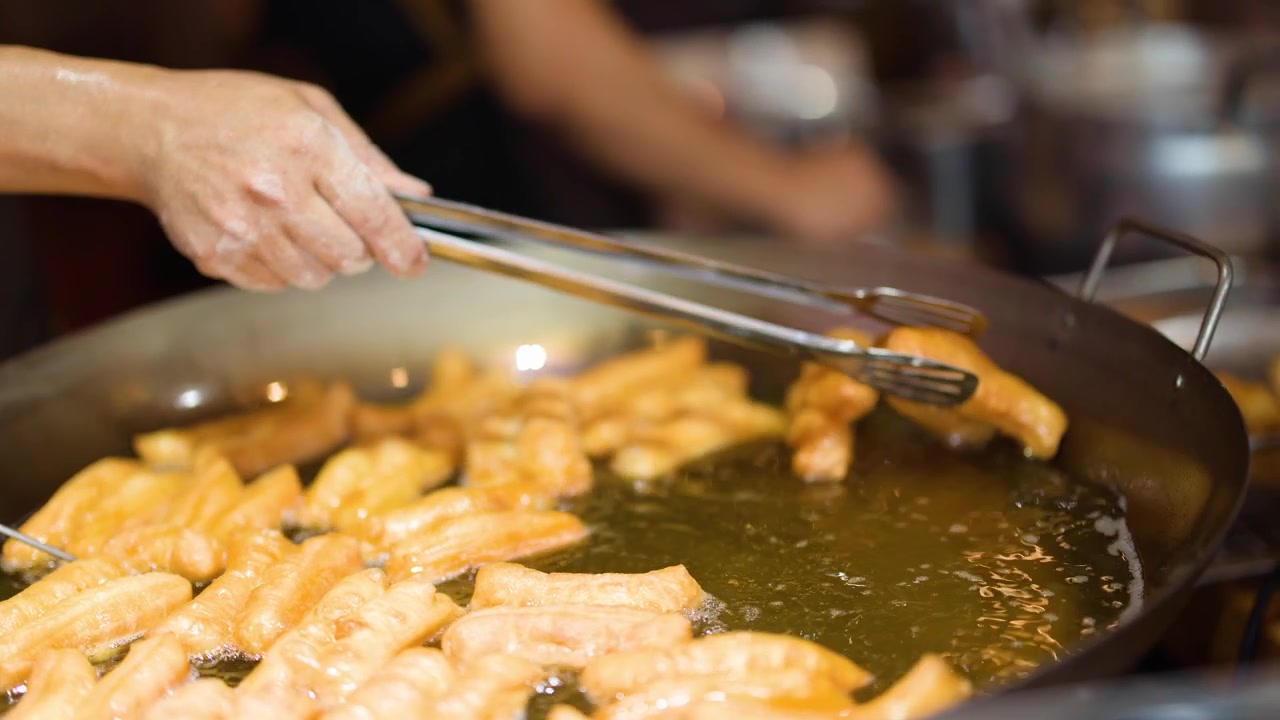 Close-up of hands using metal tongs to flip golden dough sticks in hot oil.