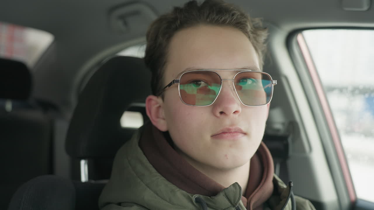 close up portrait of young boy in glasses wearing winter jacket sitting inside car, calmly staring forward with relaxed expression, interior softly lit with cold daylight and blurred urban background