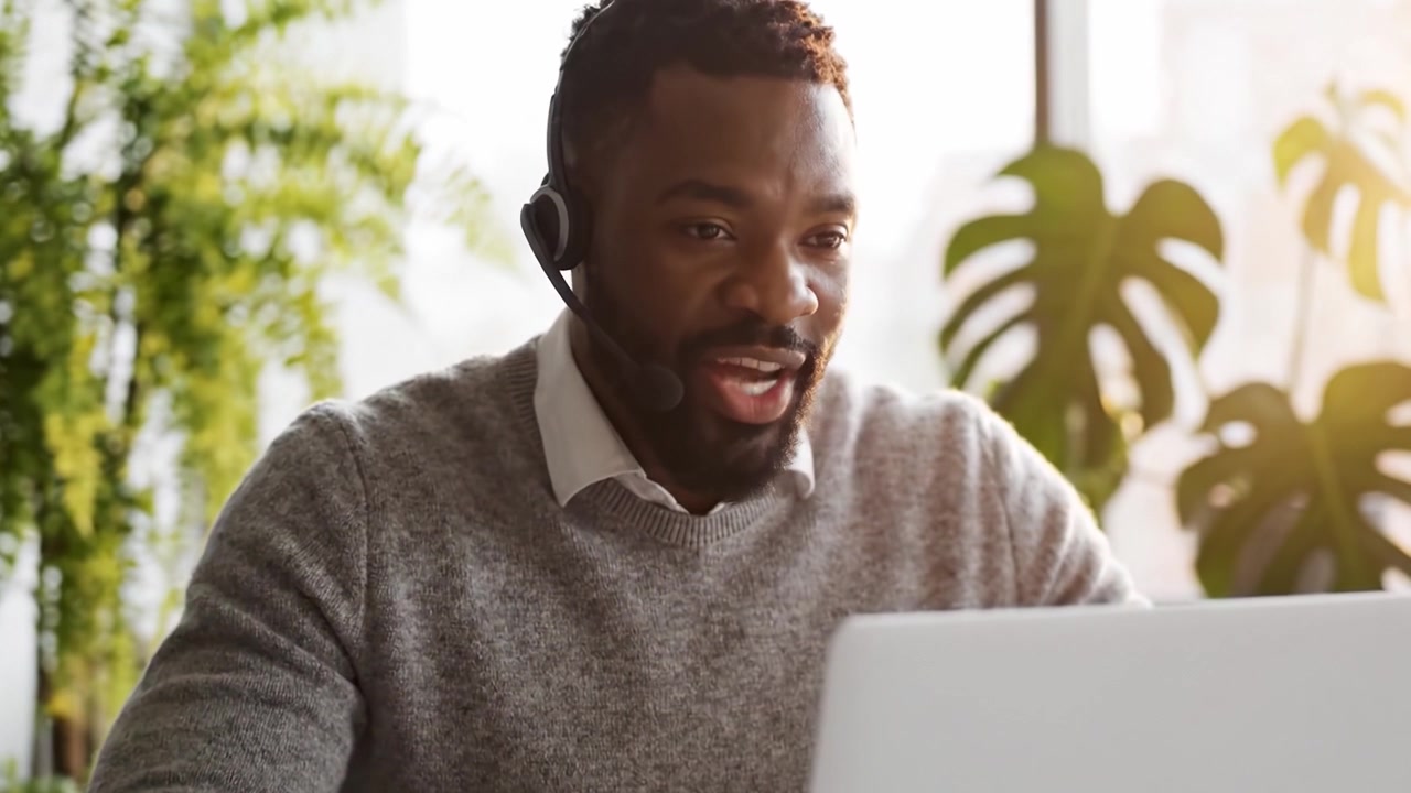 Fokuseret professionel bruger headset og bærbar computer, mens vedkommende arbejder eksternt i et lyst, grønt indendørs rum og deltager i virtuel kommunikation eller et møde.