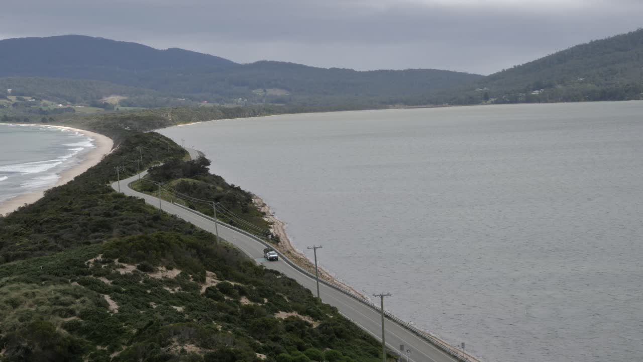 ute blanco solitario con remolque conduce por la carretera del istmo de la isla rodeada de aguas oceánicas de playa y una pequeña montaña en un día nublado y sombrío