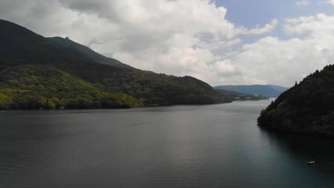 Un-Dron-Aéreo-Volador-Hacia-Adelante-Disparó-Sobre-Un-Hermoso-Lago-Con-Colinas-Forestales-Y-Un-Cielo-Azul-Y-Nublado