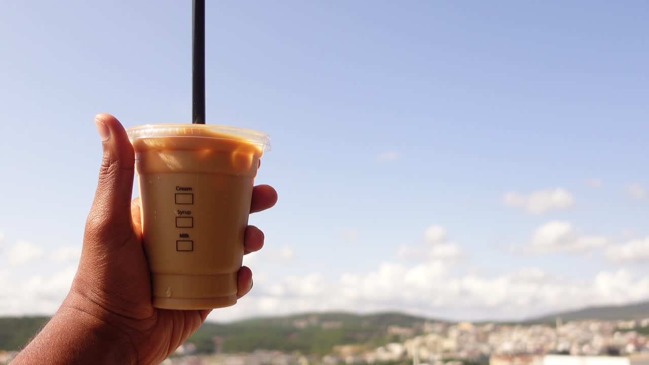 una mano sosteniendo una taza de café helado frente a una hermosa vista de una ciudad y montañas