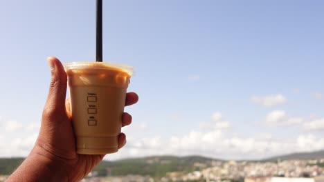 a hand holding a cup of iced coffee in front of a beautiful view of a city and mountains