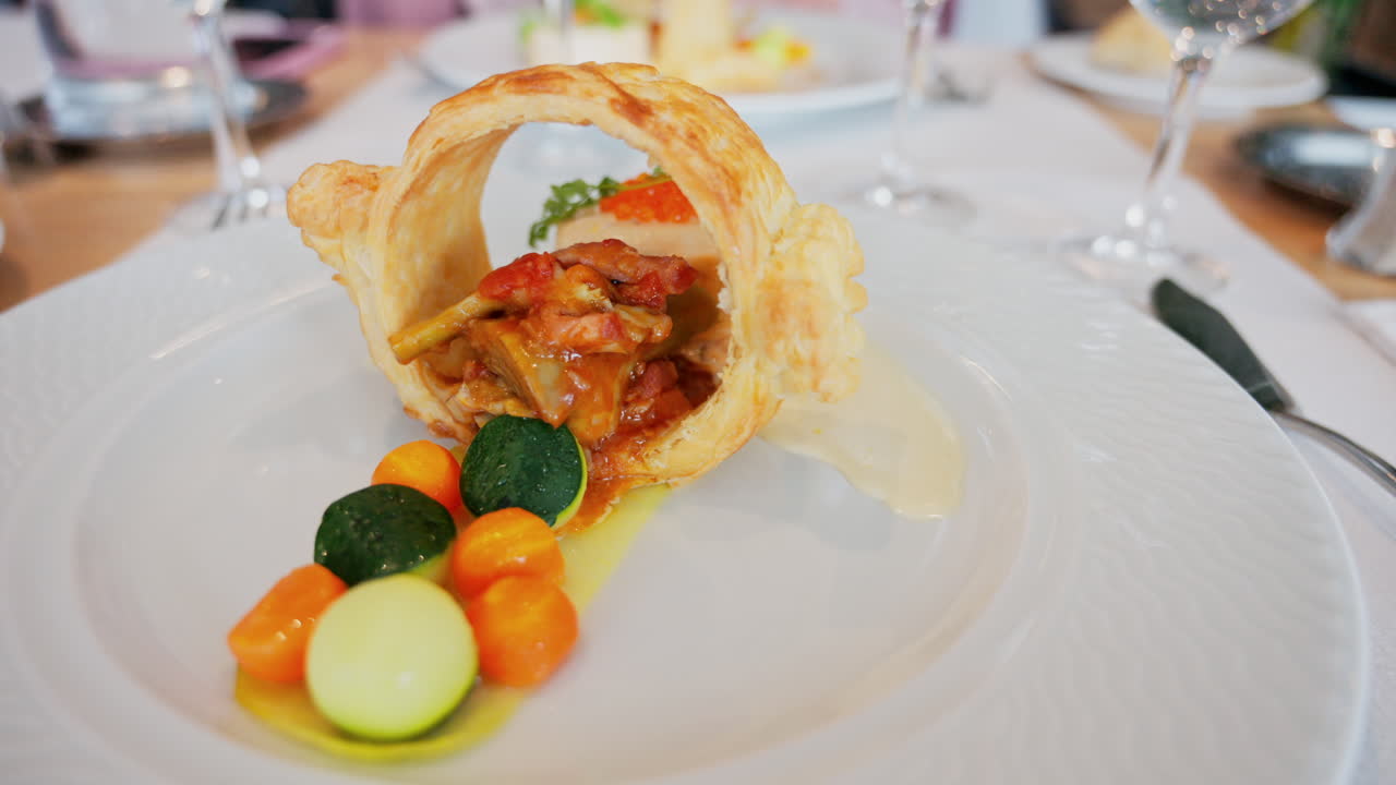 Close up of a golden, circular puff pastry ring filled with a tomato-based stew, with round vegetables and creamy white sauce on a white plate at a restaurant