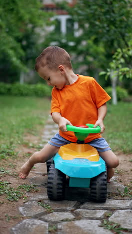 lovely kid riding his toy car by the paved path in the garden. happy kid approaches camera on his toy vehicle. vertical video.