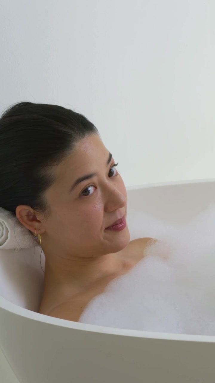 Vertical video: Settling back woman touching bubble foam for rest in freestanding tub on towel