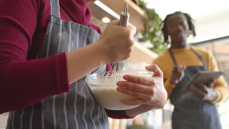 Happy-diverse-couple-in-aprons-using-tablet,-mixing-dough-and-talking-in-kitchen,-slow-motion