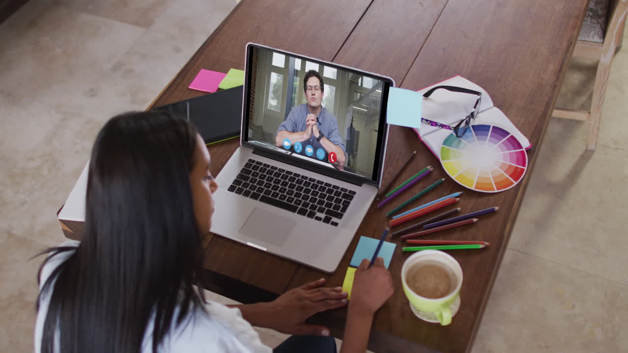 mujer caucásica usando una computadora portátil en una videollamada con un colega masculino y tomando notas trabajando desde casa