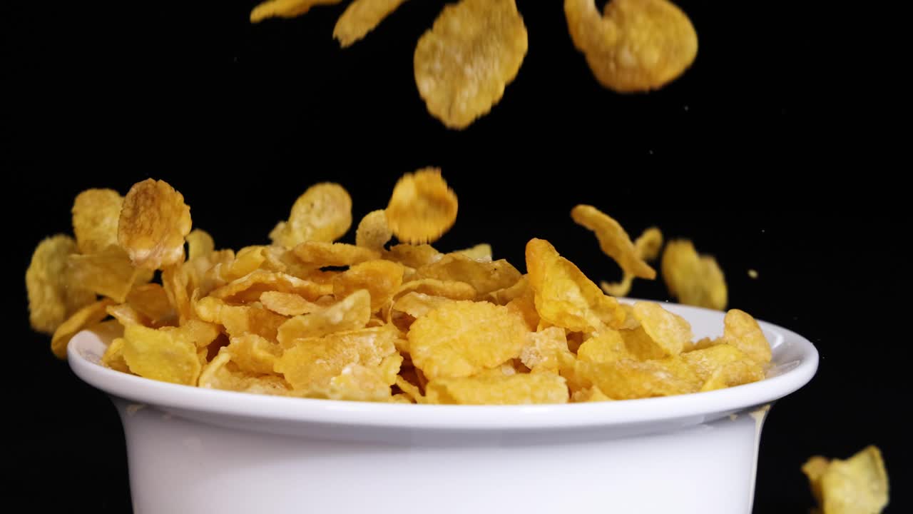 Golden corn flakes cascade into a white bowl, set against a stark black background, creating a dynamic visual contrast