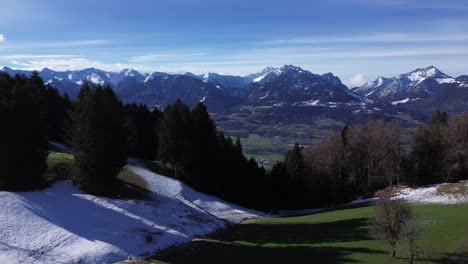 drone fly towards forest with town and winter mountain landscape with snowcapped mountains in background on a sunny day sky in austria