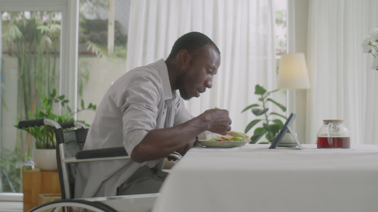 Man in Wheelchair Eating Food and Watching Video on Tablet