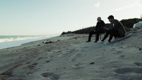 son takes elderly father to see the sea at sunset seated on the beach