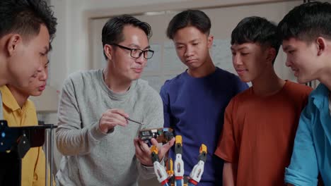 hombre asiático con una computadora portátil, impresión 3d, y una mano ciborg explicando sobre la placa de circuito a los niños en la escuela