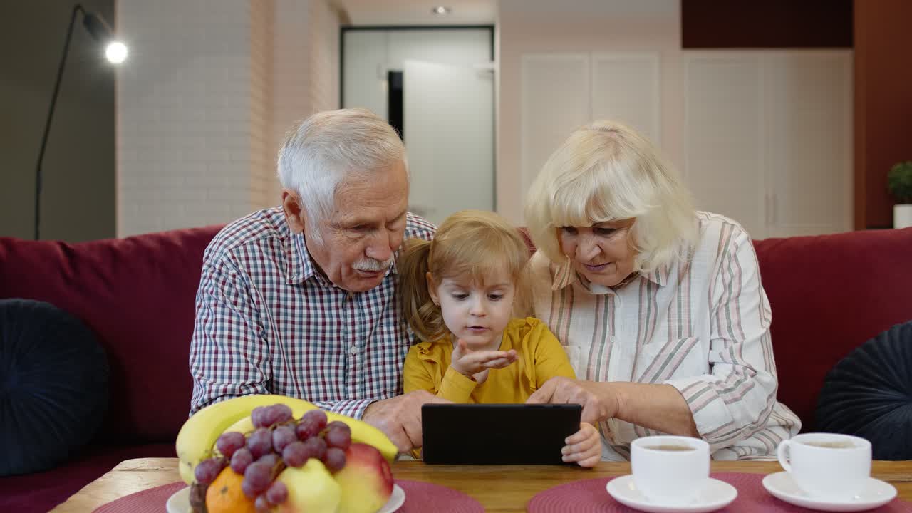 Grandfather and grandmother sitting in living room and teaching small granddaughter using tablet